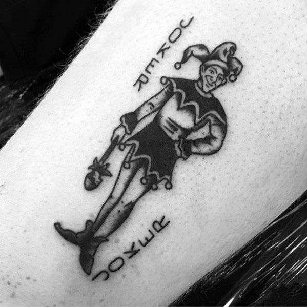 A black and gray tattoo of a jester in a playful pose, holding a scepter, with stylized lettering on either side of the figure.