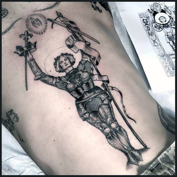 Fine line Joan of Arc rib tattoo showing her in full armor with a halo, holding a sword and flag in a triumphant pose.