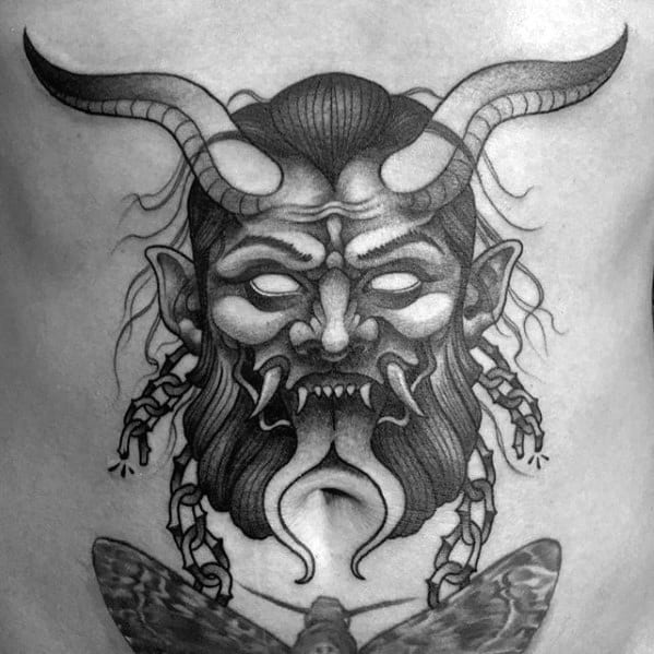 Black and grey Krampus tattoo on torso showing a horned head with glowing eyes, twisted chains, fangs, and a moth below.