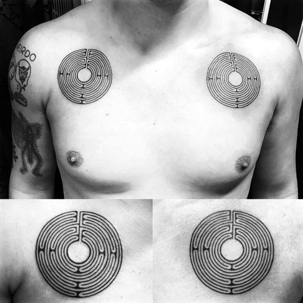Symmetrical black ink labyrinth tattoos on a man's chest, featuring two identical circular maze designs positioned above each pectoral muscle.