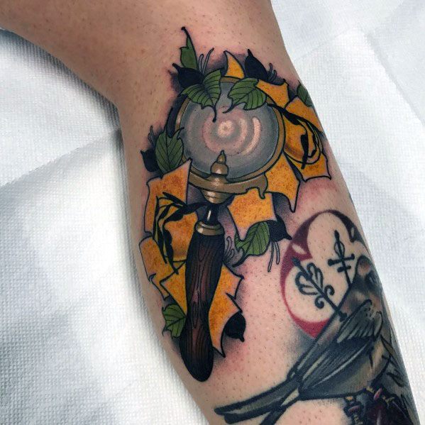 Colorful tattoo of a magnifying glass with a wooden handle, surrounded by torn parchment and green leaves, creating a bold and vintage-inspired design.