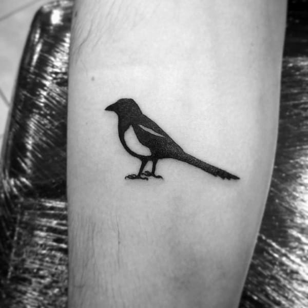 Minimalist black magpie tattoo on forearm, featuring a simple silhouette design with clean lines for a sleek and understated look.