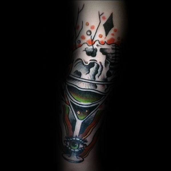 Dark-themed martini glass tattoo featuring a skull, an eye, and abstract elements. A bold, eerie design with intricate shading and orange accents.