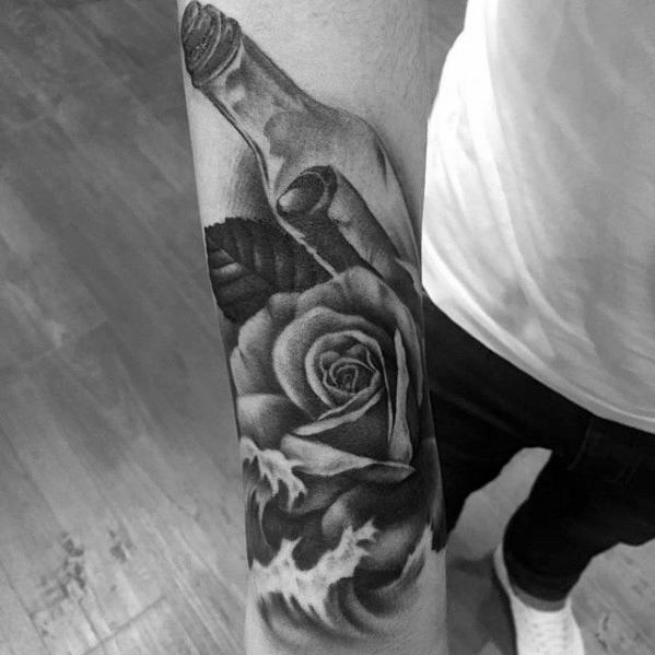Black and gray tattoo of a message in a bottle, rose, and leaves on a person's forearm