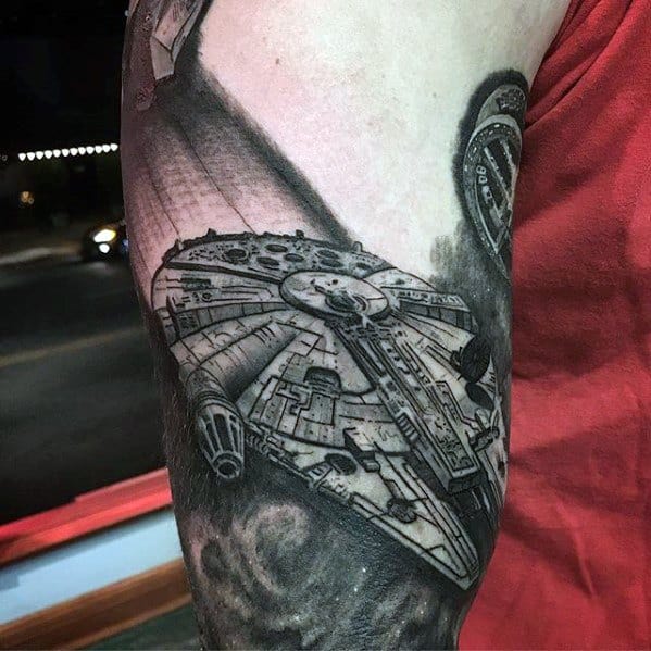 Realistic black and grey Millennium Falcon tattoo in flight, featuring dramatic shading and space-themed background on the upper arm.
