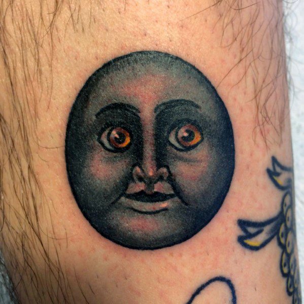 Tattoo of a black moon face with detailed shading and expressive orange eyes