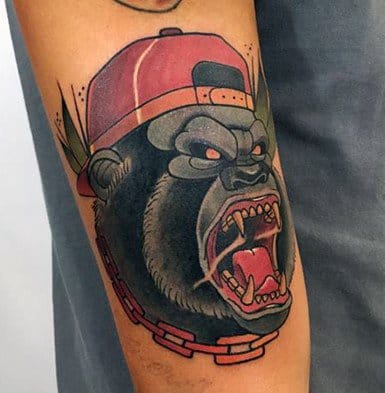 Neo-traditional gorilla tattoo wearing a red cap and chain, showing fierce expression with sharp teeth and bold outlines.