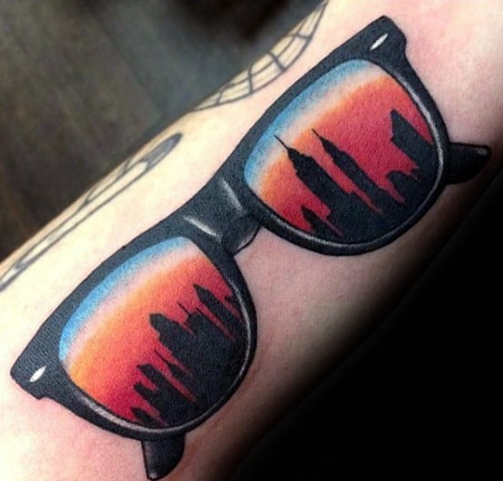 Tattoo of sunglasses reflecting a New York City skyline at sunset, with bold black outlines and vibrant orange-blue hues.