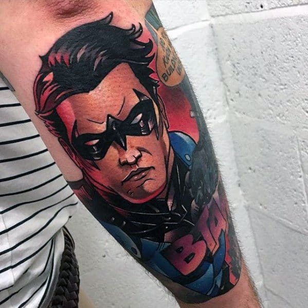 Bold comic-style Nightwing tattoo on the forearm featuring vivid colors, sharp outlines, and a speech bubble for dramatic flair.
