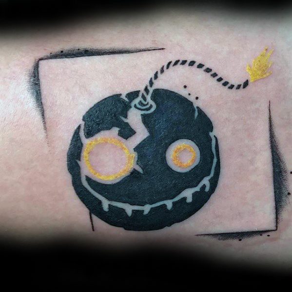 Tattoo of Junkrat’s bomb logo from Overwatch, featuring a cracked smiley face design with a lit fuse, framed in black ink.