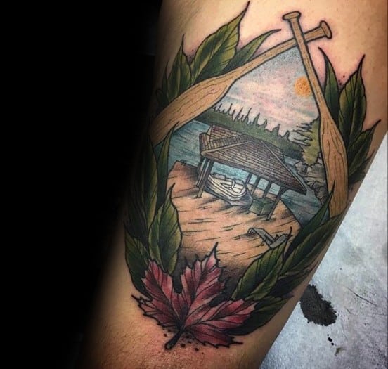 Colorful tattoo featuring crossed paddles framing a serene lakeside scene with a dock, canoe, and maple leaf, symbolizing nature and adventure.