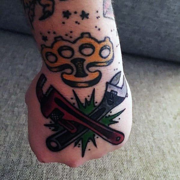 Tattoo on the hand showing crossed pipe and adjustable wrenches over a green burst, with brass knuckles and stars above.