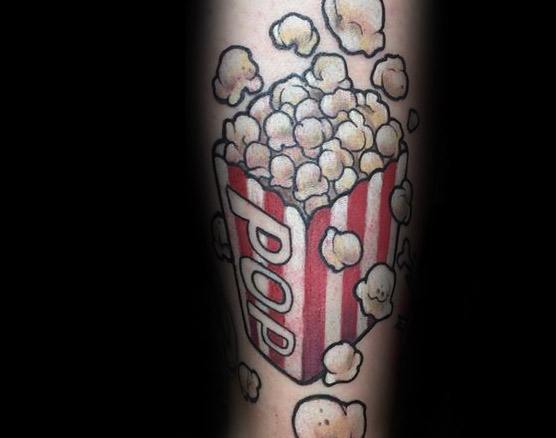 Tattoo of a red-striped popcorn box with the word POP and smiling kernels floating around in a playful, cartoonish style.