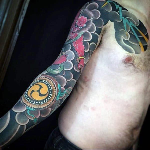 Minimalist Raijin full sleeve tattoo with bold black clouds, red thunder god details, and golden tomoe drum symbol on the forearm.