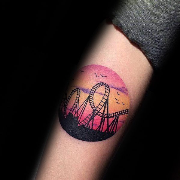 Vibrant roller coaster tattoo with silhouette loops set against a round sunset sky in pink, orange, and purple tones.