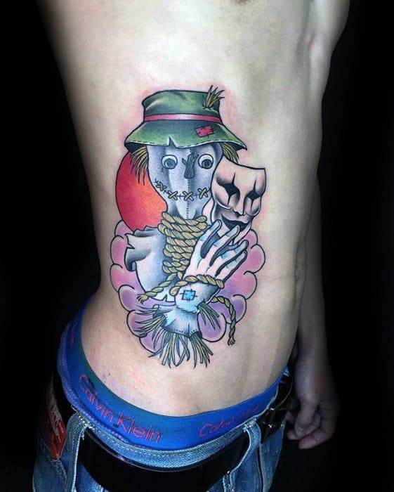 Colorful scarecrow tattoo on the ribcage featuring a stitched face, green hat, red sun, rope neck, and a dramatic hand holding a mask.