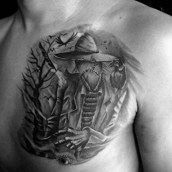 Black and grey scarecrow chest tattoo with stitched mouth, wide-brim hat, perched crow, eerie trees, and flying birds in the background.
