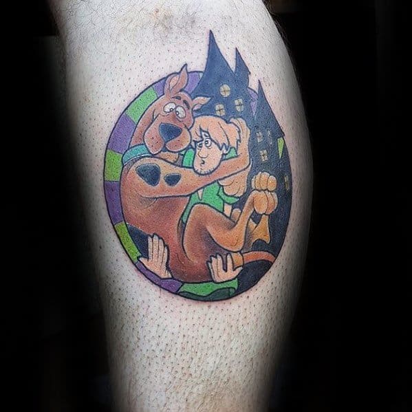 Tattoo of a frightened Scooby-Doo clinging to Shaggy with a purple and green border and spooky houses in the background.