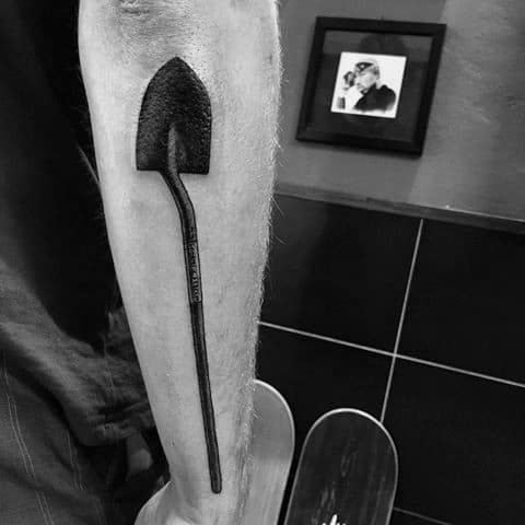 Bold black shovel tattoo on the forearm with a long, slightly curved handle and a darkened spade, creating a strong and striking design.