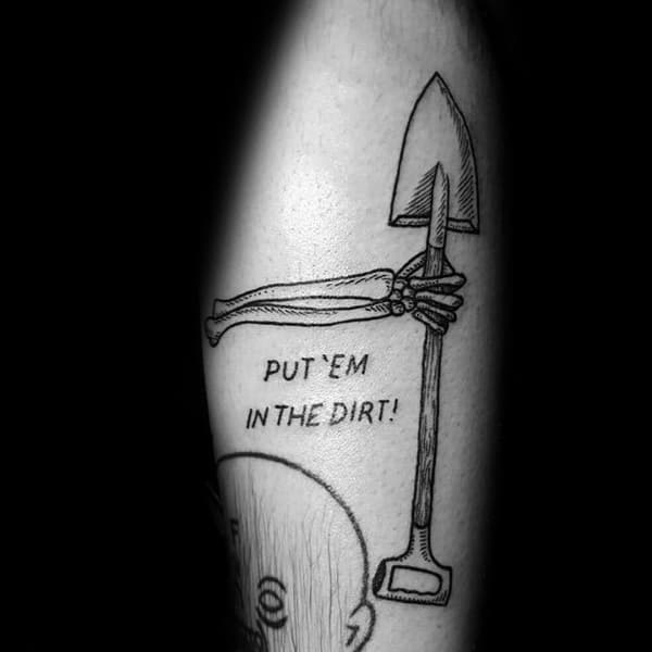 Black and grey tattoo of a skeletal hand gripping a shovel, with the phrase "PUT 'EM IN THE DIRT!" adding a bold and edgy statement.