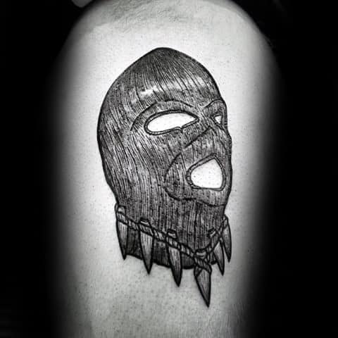 Tattoo of a textured ski mask with open eyes and mouth, featuring a necklace of sharp teeth or claws at the base.