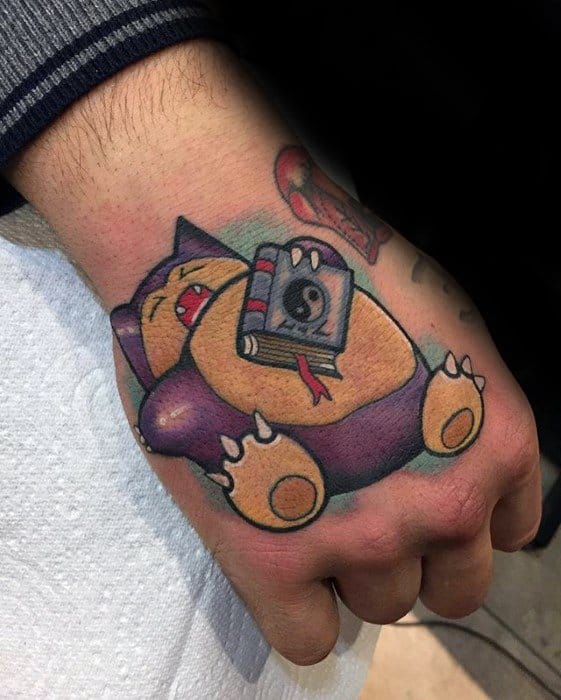 Colorful Snorlax tattoo on hand, sleeping peacefully while holding a book with a yin-yang symbol on the cover.