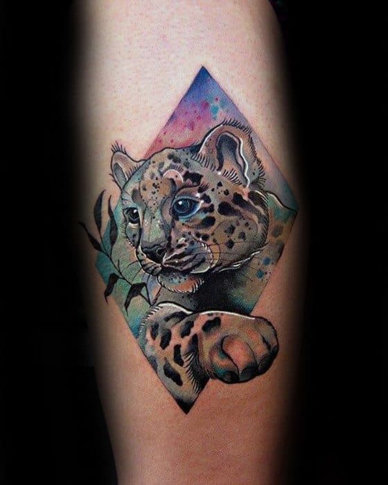 Colorful snow leopard cub tattoo with a raised paw, framed in a diamond shape and accented with leaves and watercolor splashes.