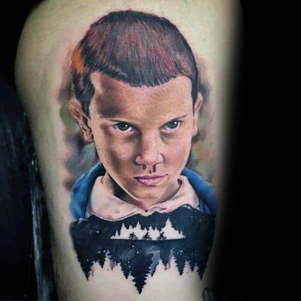 Realistic tattoo of Eleven from Stranger Things with a focused expression, above a silhouette of trees and the Upside Down reflection.