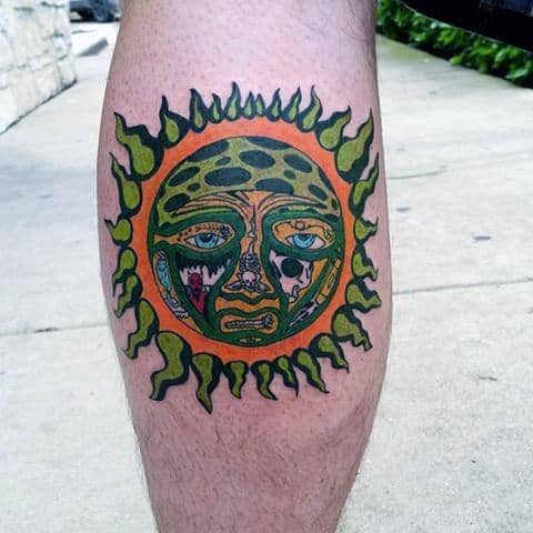 Sublime sun face tattoo on the calf, featuring vivid green and orange tones with expressive eyes and flame-like border.