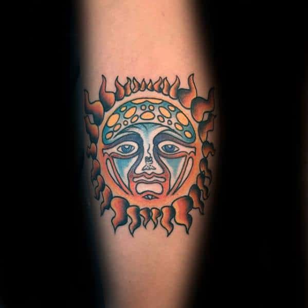 Minimalist Sublime sun face tattoo with blue and orange tones, bold outlines, and simplified flame rays on the forearm.