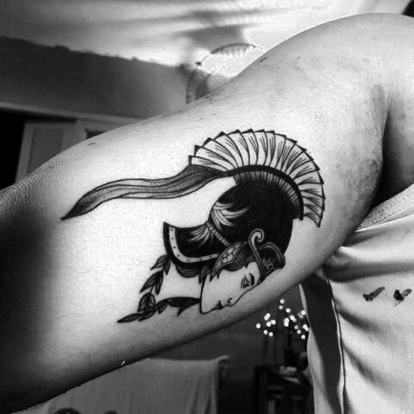 Small Athena tattoo on bicep with bold black lines, plumed helmet, and downward gaze in a minimalist illustrative style