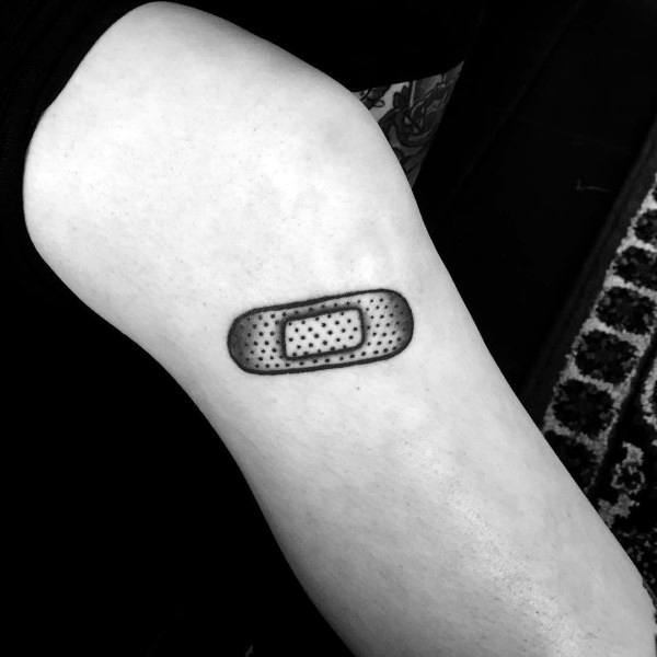 Small black and grey Band-Aid tattoo with detailed dotwork shading, placed neatly on the upper arm for a clean, subtle look.