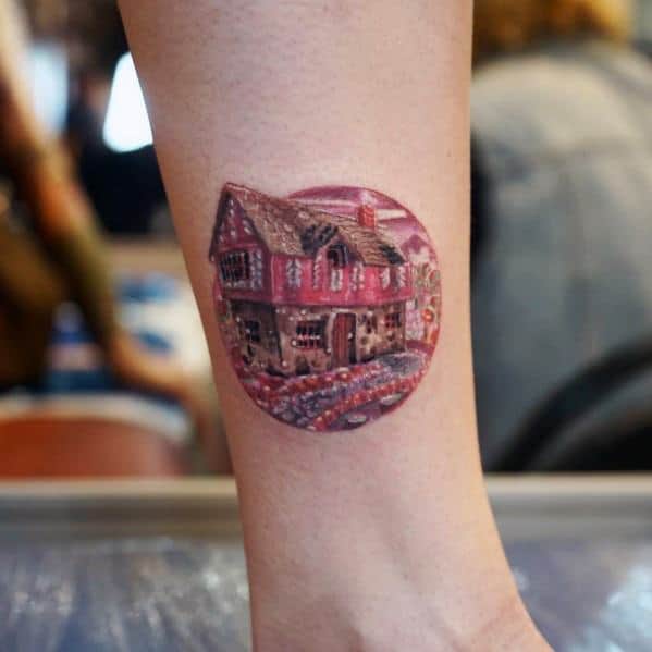 Detailed candy-themed tattoo of a gingerbread house in a circular frame, featuring rich textures and warm tones for a storybook-like effect.