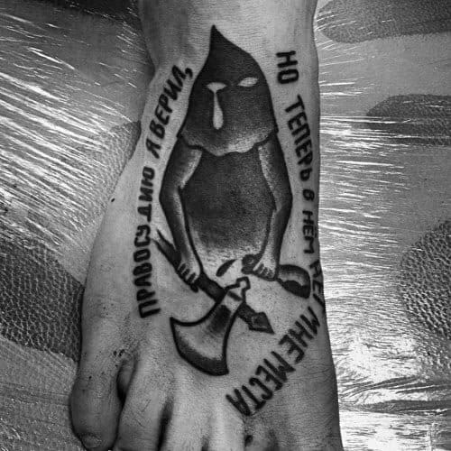 Black and grey executioner tattoo on foot, featuring a hooded figure holding two axes, surrounded by Russian text in bold shading.
