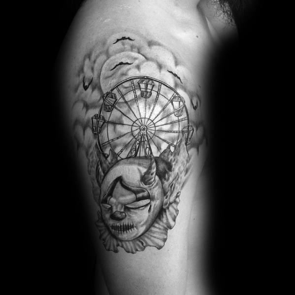 Dark-themed Ferris wheel tattoo with eerie clouds, bats, and a sinister clown mask, creating a haunting carnival-inspired design on the upper arm.