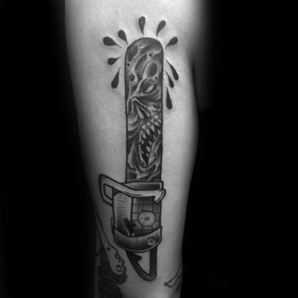Black and grey Evil Dead tattoo of a chainsaw with a terrifying deadite face carved into the blade, dripping with dark ink splatters.