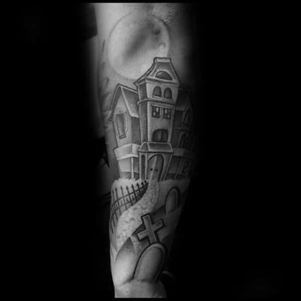 Forearm tattoo of a haunted mansion on a hill with a winding path, graveyard crosses, iron fence, and a full moon above.