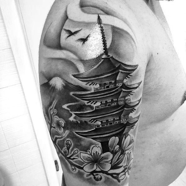 Black and grey upper arm tattoo of a Japanese pagoda with cherry blossoms, flying birds, Mount Fuji, and swirling clouds.