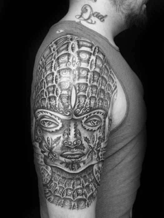Large black and grey Tool-inspired upper arm tattoo of a symmetrical face with ornate patterns, repeated textures, and deep shading.