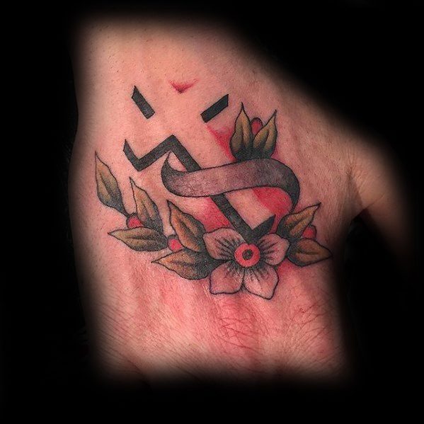 Small black cross tattoo on the hand with a red-pink background, wrapped in a ribbon and surrounded by leaves and a flower.
