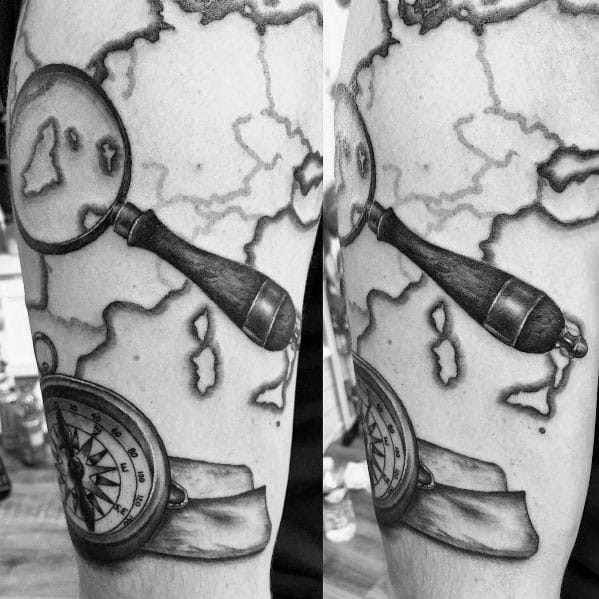 Tattoo featuring a magnifying glass over a world map with a compass and scroll, creating a detailed black and gray exploration-themed design.