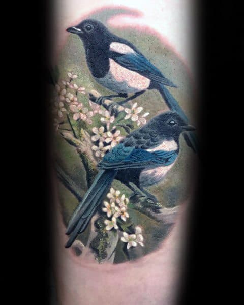 Realistic magpie tattoo featuring two birds perched on a flowering branch, with intricate details and soft shading for a natural, lifelike design.