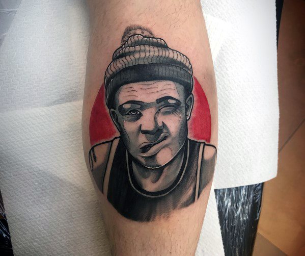 Calf tattoo featuring a bold black and grey portrait of a beanie-wearing figure with a playful expression, set against a red circular backdrop.