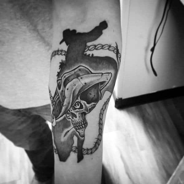 Black and grey tattoo featuring a cowboy silhouette riding a bull, with a detailed skull wearing a cowboy hat and a rope framing the design.