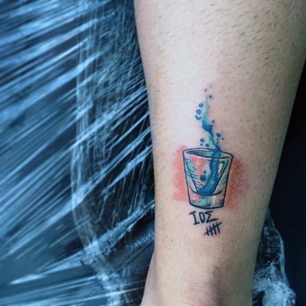 Small shot glass tattoo with a blue liquid splash, bubbles, and "1oz" with tally marks below, creating a vibrant and dynamic design.