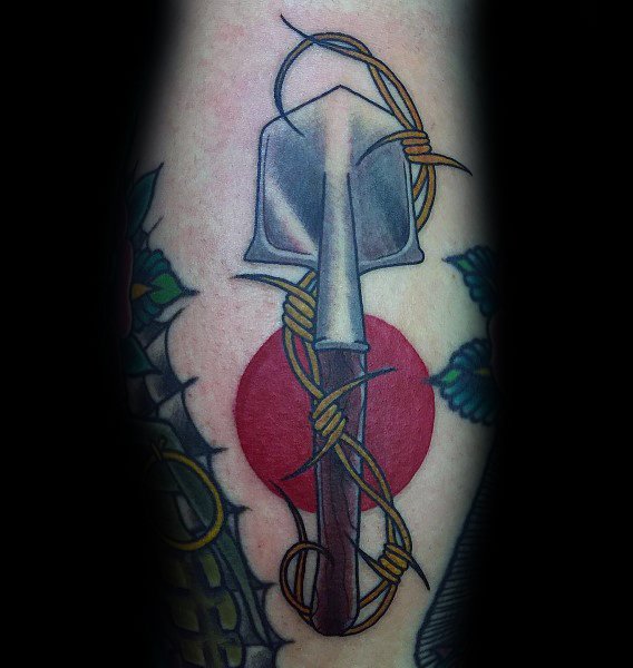 Shovel tattoo with a red sun in the background, wrapped in barbed wire, symbolizing resilience, struggle, and strength in a bold design.
