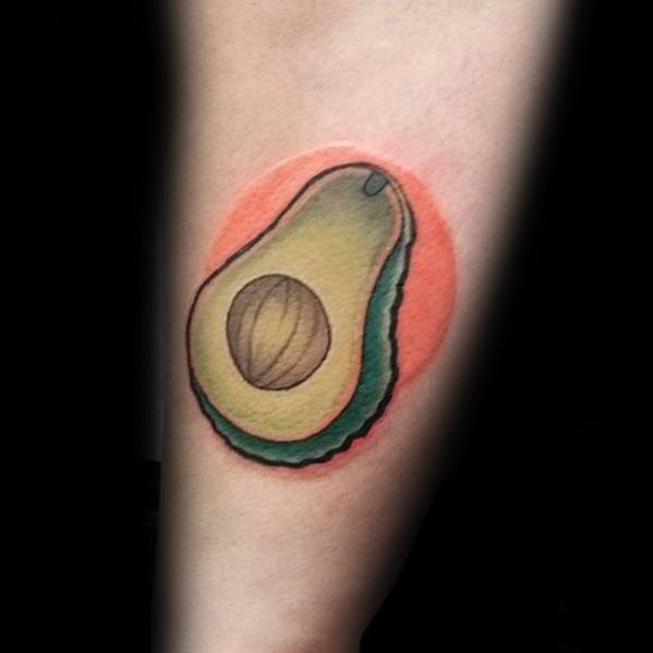 Softly shaded avocado tattoo with a warm orange background circle, blending pastel tones for a gentle, artistic look on the forearm.