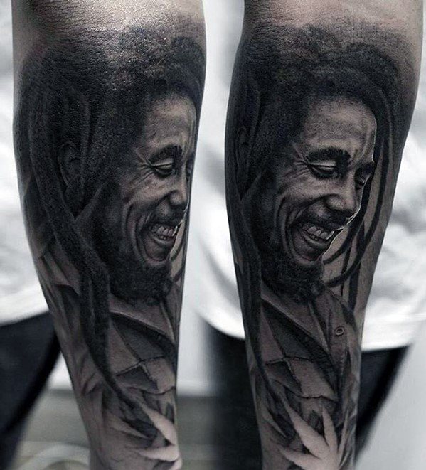 Black and gray tattoo of Bob Marley smiling with detailed shading, dreadlocks, and a subtle cannabis leaf design at the bottom.