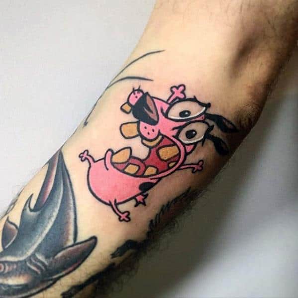 Tattoo of Courage the Cowardly Dog in a dramatic, scared pose with bulging eyes and open mouth, inked on the inner arm.
