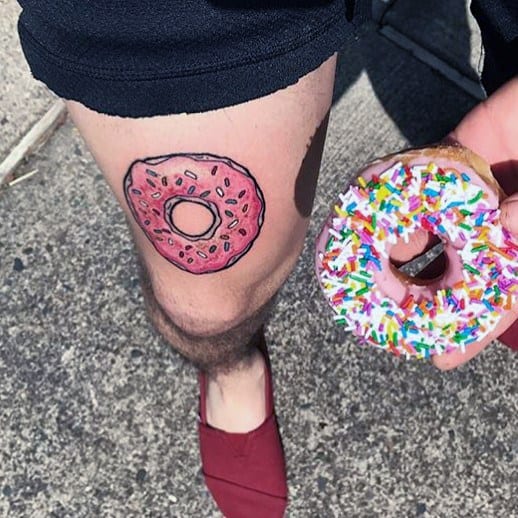 Tattoo of a pink frosted donut with sprinkles on the thigh, humorously mirrored by a real sprinkled donut held beside it.